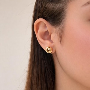 Peut inclure: Une paire de boucles d'oreilles à clous dorées. Chaque boucle d'oreille présente un motif de nœud texturé. Les boucles d'oreilles sont rondes et ont une finition polie, réfléchissant la lumière. Les boucles d'oreilles sont portées.