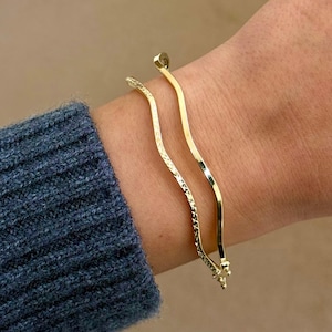 May include: Two gold-colored bracelets with a wavy design. One bracelet has a textured pattern, while the other is smooth. The bracelets are worn on a wrist, with a blue sweater sleeve in the background. The bracelets are a jewelry item.