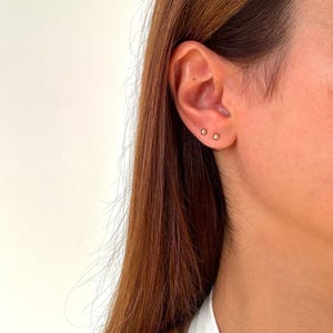 Peut inclure: Trois petites boucles d'oreilles à clous dorées sont visibles sur une oreille. Les boucles d'oreilles sont espacées uniformément et semblent rondes. L'oreille est placée sur un fond de longs cheveux bruns et d'une chemise blanche.