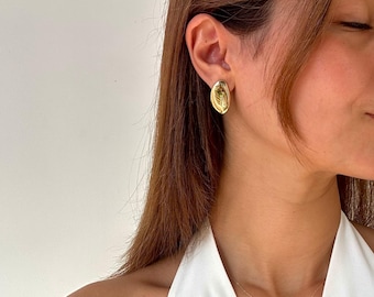 May include: Gold-toned oval stud earrings. Each earring features a textured design within the oval shape. The earrings are worn on the ear of a person wearing a white top and a gold necklace.
