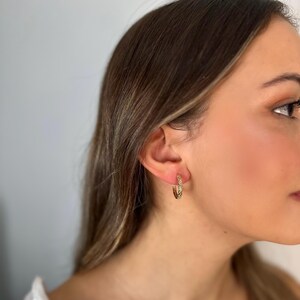Peut inclure: Gros plan sur l'oreille d'une personne portant une paire de créoles dorées. Les boucles d'oreilles sont texturées et ont une forme légèrement ovale.