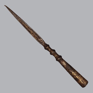 May include: A long, slender metal letter opener with a dark, weathered finish and intricate gold-colored filigree engravings. The handle features a textured, cross-hatched pattern and decorative ridges, while the blade tapers to a sharp point.