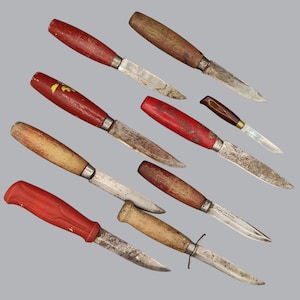 Op de afbeelding: Een verzameling van acht messen met een vaststaand lemmet en diverse handgrepen, waaronder rood gelakt hout, natuurlijk hout en gestructureerd rood kunststof. De metalen lemmeten vertonen tekenen van slijtage, oxidatie en roest.