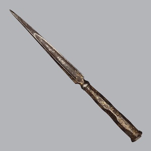 Op de afbeelding: Een lange, slanke metalen briefopener met een puntig, donkergrijs lemmet en een cilindervormig handvat. Het handvat en de basis van het lemmet zijn voorzien van ingewikkelde, versleten goudkleurige filigraanpatronen in het donkere metaal.