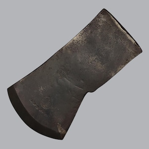 May include: A dark gray metal axe head with a curved cutting edge and a weathered, textured surface. The metal shows signs of age and use, with a visible stamped mark near the blade edge. The design features a wide, flat poll and a tapered bit.