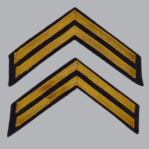 May include: Two identical chevron-shaped patches featuring a dark navy blue fabric base with two parallel, raised gold-colored embroidered stripes running along the center of each chevron, creating a distinct V-shaped military-style insignia.
