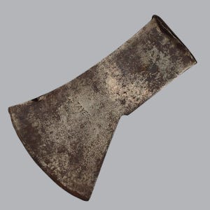 May include: A weathered metal axe head with a dark, mottled patina and a curved cutting edge. The word STAYER is stamped clearly into the center of the metal surface. The tool features a flared blade design and a rectangular opening for a handle.