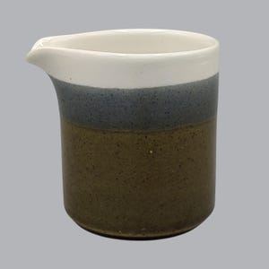 May include: A ceramic creamer pitcher featuring a cylindrical body with a pour spout. The exterior displays a tri-color design with a white upper section, a speckled grey middle band, and a speckled brown base, all with a smooth finish.
