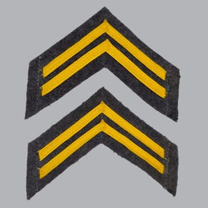 May include: Two dark gray felt chevron patches, each featuring two parallel embroidered yellow stripes arranged in a V-shape. These military-style rank insignia patches have straight, clean edges and are positioned one above the other.