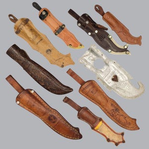 Op de afbeelding: Een verzameling van acht messenscheden, waaronder zeven gemaakt van verschillende tinten bruin en beige leer met decoratieve stiksels, reliëfpatronen en metalen klinknagels, naast één zilverkleurige metalen schede met een hartvormige uitsparing.