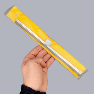 Op de afbeelding: Een zilveren metalen gereedschap, mogelijk een liniaal of een rechte hoek, is verpakt in een heldergele plastic hoes. Het gereedschap wordt in de hand van een persoon gehouden tegen een grijze achtergrond. Een kleine, lichtblauwe sticker zit op de verpakking.