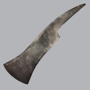 Op de afbeelding: Een donkergrijs, verweerd metalen gereedschap met een brede, platte snijkant en een puntig, gebogen uiteinde. Het oppervlak vertoont zichtbare krassen en een gestempelde inscriptie met de tekst "WETTI" boven "ASTORUKS". Het gereedschap heeft een robuuste uitstraling.