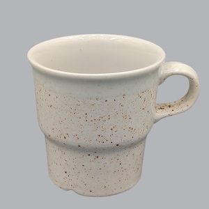 May include: A white ceramic mug with a speckled design. The mug has a cylindrical shape with a handle and a stepped base. The speckled pattern is a mix of brown and tan dots. The interior of the mug is white.