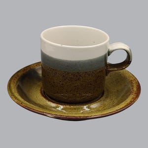 Op de afbeelding: Een keramische koffiekop en schotel set. De beker heeft een witte binnenkant, een grijze band en een bruine getextureerde basis. De schotel is bijpassend olijfgroen met een bruine rand. De set is ontworpen voor het serveren van warme dranken.