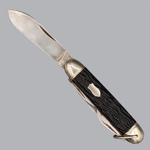 May include: A folding pocket knife with a single extended silver-toned steel blade and a textured black handle. The handle features a small silver-toned shield-shaped inlay and polished metal bolsters at both ends, with a small metal loop at the base.
