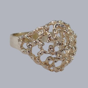 May include: A gold-colored ring with a unique, openwork design. The ring's surface has a textured, organic appearance, with numerous small holes and irregular shapes. The band is smooth and solid, contrasting with the intricate top. A statement piece of jewelry.
