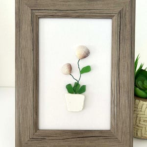 Könnte beinhalten: Gerahmtes Kunstwerk mit einem Blumengesteck aus Muscheln. Das Kunstwerk zeigt zwei Muscheln als Blüten, grüne Glasblätter und einen weißen Topf. Der Rahmen hat eine rustikale grau-braune Farbe.