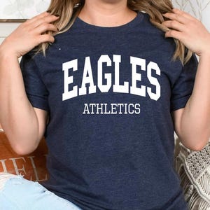 May include: A dark navy blue short-sleeved t-shirt featuring the white text EAGLES in a bold arched collegiate font with the word ATHLETICS printed in a smaller white sans-serif font directly underneath the main text on the chest area.