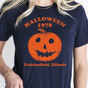 May include: A navy blue t-shirt with a graphic of an orange pumpkin with a scary face. The text "HALLOWEEN 1978 Haddonfield, Illinois" is printed on the shirt.