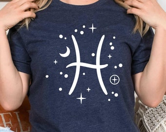 May include: A navy blue t-shirt with a white zodiac symbol for Pisces, surrounded by stars and a crescent moon. The shirt is being worn by a person sitting on a bench with pillows.