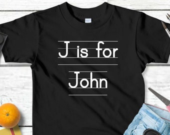 May include: Black t-shirt with white text that says "J is for John" on a white lined background. The shirt is laid out on a white wooden surface with school supplies around it.