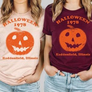 May include: Two women wearing t-shirts with a vintage Halloween design. The shirts are peach and maroon and feature a pumpkin with a face and the text "Halloween 1978 Haddonfield, Illinois".