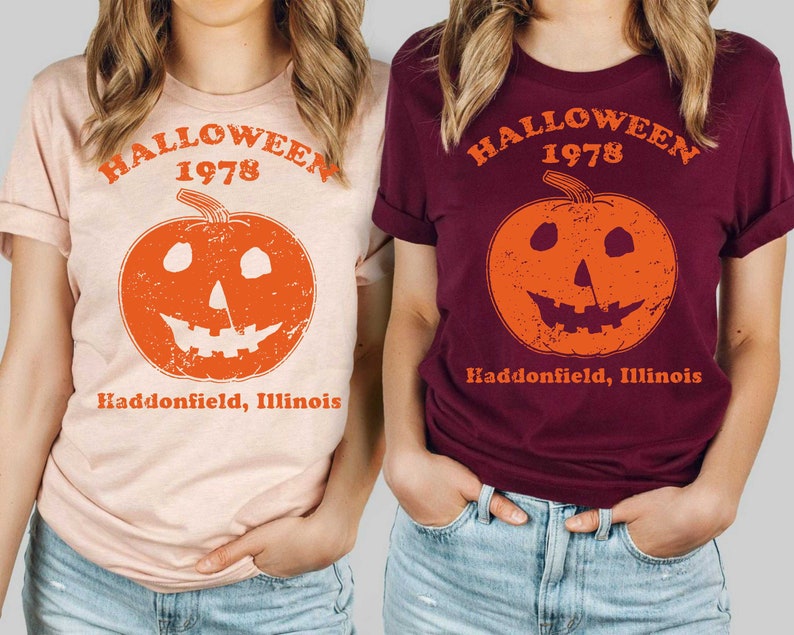 May include: Two women wearing t-shirts with a vintage Halloween design. The shirts are peach and maroon and feature a pumpkin with a face and the text "Halloween 1978 Haddonfield, Illinois".