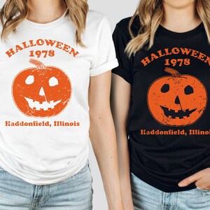 May include: Two women wearing t-shirts with a vintage Halloween graphic. The graphic features a jack-o'-lantern with a distressed look and the text "Halloween 1978 Haddonfield, Illinois".