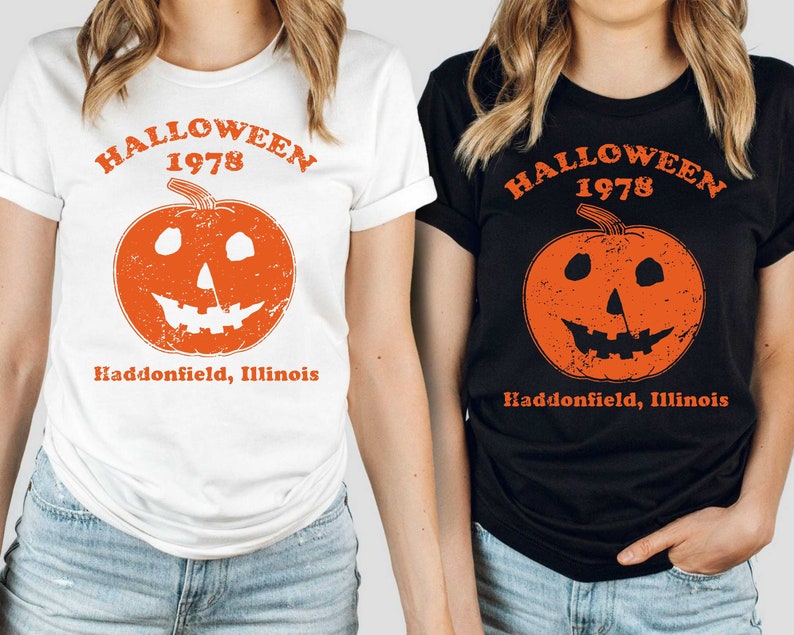 May include: Two women wearing t-shirts with a vintage Halloween graphic. The graphic features a jack-o'-lantern with a distressed look and the text "Halloween 1978 Haddonfield, Illinois".