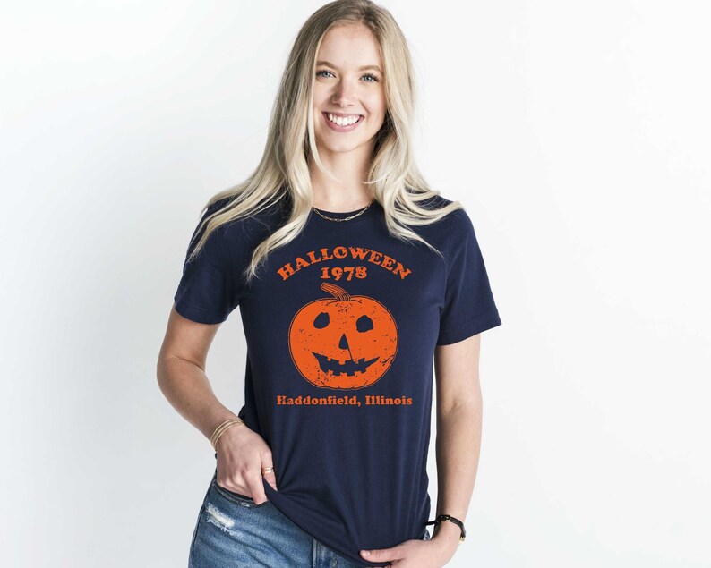 May include: A navy blue t-shirt with a graphic of an orange pumpkin with a scary face. The text "HALLOWEEN 1978 Haddonfield, Illinois" is printed on the shirt.