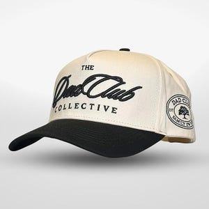 May include: A white and black baseball cap with the text "The Dad Club Collective" in black cursive script on the front. The back of the cap has a black and white logo with the text "Dad Club Est. Family Owned".