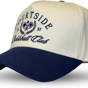 May include: A white and navy blue baseball cap with the text "Courtside Pickleball Club" embroidered on the front. The logo features a wreath with two pickleball paddles inside.