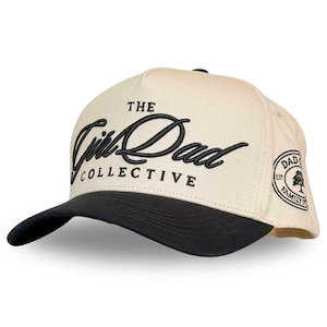 May include: A cream-colored baseball cap with a black brim. The cap features the embroidered text "THE Girl Dad COLLECTIVE" in black script. A circular patch on the side reads "DAD FAMILY CO." with a tree graphic.