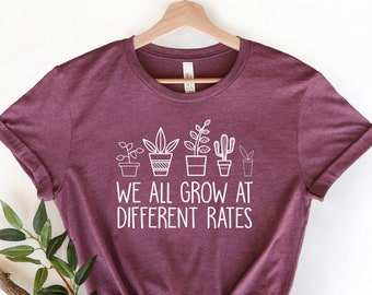 May include: A maroon t-shirt with a white graphic of potted plants and the text "We all grow at different rates".