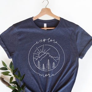 May include: A navy blue t-shirt with a white graphic of a mountain range, sun, and trees inside a circle. The text "explore more" is written above and below the graphic.
