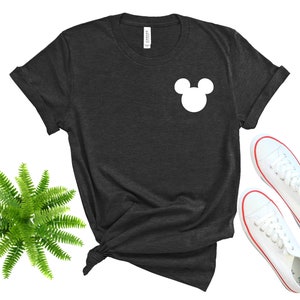 May include: A dark grey t-shirt with a white Mickey Mouse silhouette on the front. The shirt is laid flat on a white background with a pair of white sneakers.
