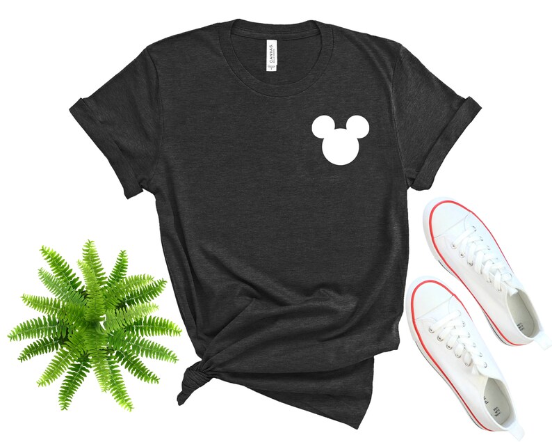 May include: A dark grey t-shirt with a white Mickey Mouse silhouette on the front. The shirt is laid flat on a white background with a pair of white sneakers.