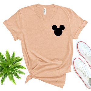 May include: A peach-colored t-shirt with a black Mickey Mouse silhouette on the front. The shirt is laid flat on a white background with a green fern plant and white sneakers.