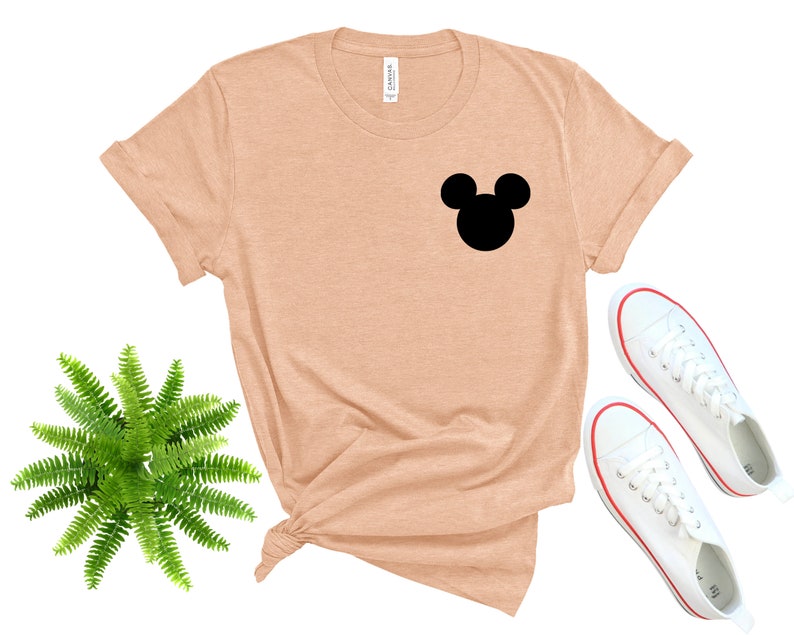 May include: A peach-colored t-shirt with a black Mickey Mouse silhouette on the front. The shirt is laid flat on a white background with a green fern plant and white sneakers.