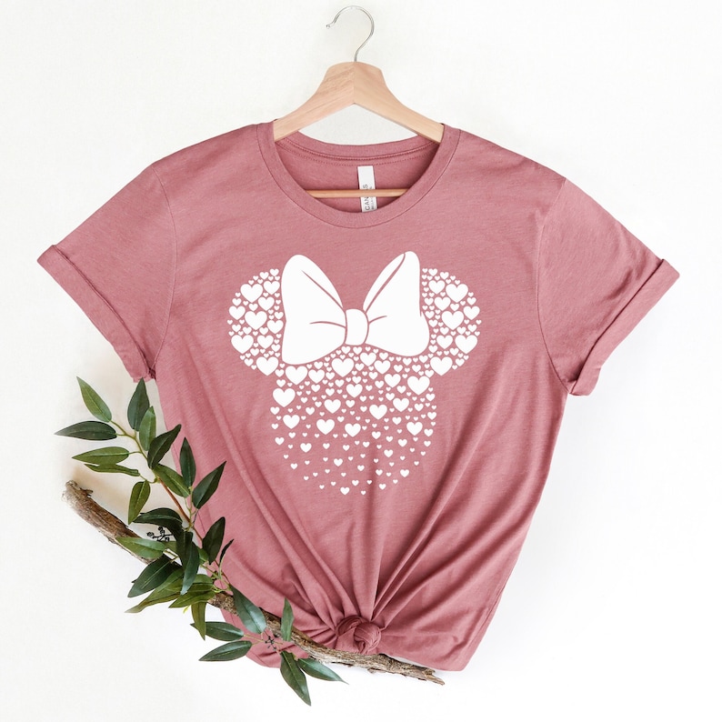 Może przedstawiać: Koszulka w kolorze malwy z białym nadrukiem głowy Myszki Minnie wykonanej z serc. Kokardka r&oacute;wnież jest wykonana z serc.