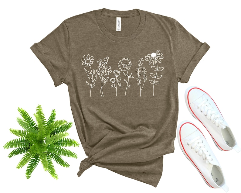 May include: A brown t-shirt with a white outline of a bouquet of flowers. The shirt is shown on a white background with a fern plant and two white sneakers with red trim.