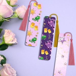 Peut inclure: Trois marque-pages colorés avec des glands. Le premier marque-page présente un fond rose avec un motif répété d'un soleil, de lanternes et d'un caméléon vert. Le deuxième marque-page présente un fond violet avec un motif répété d'une robe violette, de lanternes et d'un caméléon vert. Le troisième marque-page présente un fond rose avec un motif répété d'étoiles dorées et d'une étoile dorée avec un contour blanc.