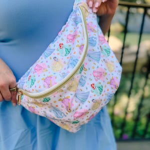 May include: A white fanny pack featuring a repeating pattern of various cartoon princesses in colorful dresses, accented with small yellow flowers. The bag includes a gold-toned zipper closure and an adjustable white strap for secure carrying.