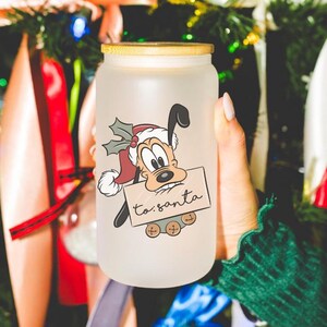 May include: A frosted glass jar with a wooden lid, featuring a cartoon dog wearing a Santa hat and holding a sign that says "to: santa". The jar is surrounded by Christmas decorations, including red and green ribbons, white bows, and a blue ribbon.