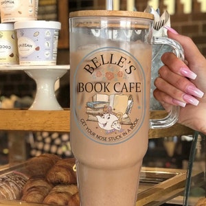 May include: A clear glass tumbler with a brown lid and a straw. The tumbler has a graphic of Belle's Book Cafe with a cartoon image of Mrs. Potts and Chip. The text reads "Belle's Book Cafe" and "Get your nose stuck in a book".