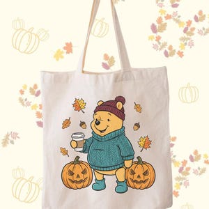May include: A cream-colored tote bag featuring a cartoon bear in a teal sweater, burgundy hat, and blue boots, holding a coffee cup. Two carved pumpkins and autumn leaves surround the bear. The bag has a simple design with long handles.