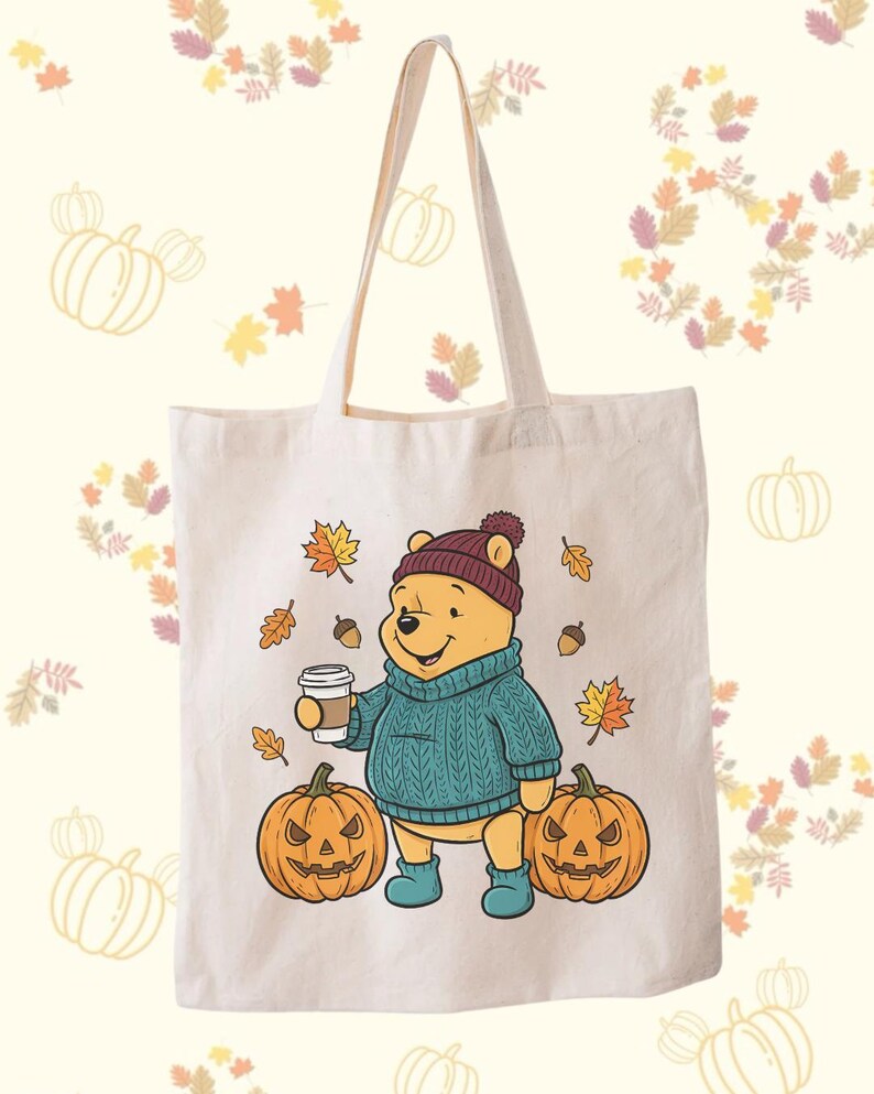 May include: A cream-colored tote bag featuring a cartoon bear in a teal sweater, burgundy hat, and blue boots, holding a coffee cup. Two carved pumpkins and autumn leaves surround the bear. The bag has a simple design with long handles.