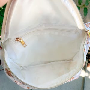 May include: Open view of a white backpack interior with a gold zipper and a small zippered pocket. The interior is lined with white fabric. The exterior of the backpack has a light beige background with a pattern of brown and pink illustrations.