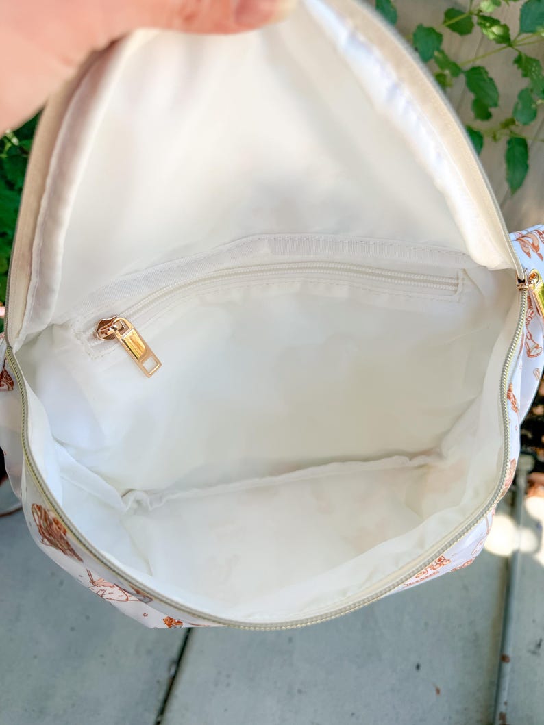 May include: Open view of a white backpack interior with a gold zipper and a small zippered pocket. The interior is lined with white fabric. The exterior of the backpack has a light beige background with a pattern of brown and pink illustrations.