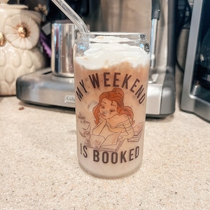 May include: A clear glass with a straw, filled with a light brown beverage topped with whipped cream. The glass has a graphic of a cartoon woman reading a book with the text "My Weekend Is Booked".